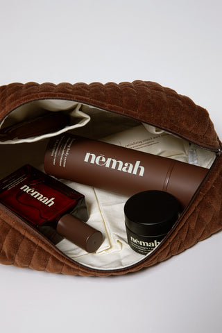 Brown quilted pouch unzipped to reveal three skincare products labeled 'nēmah': a firming body serum, a red bottle, and a small black jar with text 'nēmah' and 'nipple + lip'.
