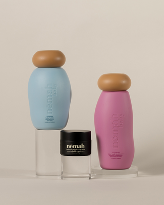 Three baby care products from nēmah baby: a blue bottle labeled 'soothing baby lotion 9.8 fl. oz. | 290 mL', a pink bottle labeled 'nourishing baby wash & shampoo 9.8 fl. oz. | 290 mL', and a black jar labeled 'renewing nipple + lip balm net wt. 1 oz | 30 g'.