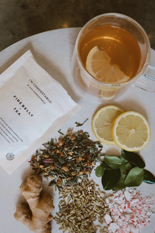 Clear glass cup of tea with a lemon slice, surrounded by two lemon slices, fresh green leaves, ginger root, fennel seeds, crushed peppermint, and a white compostable Firebelly Tea package.