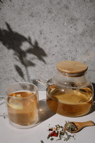 Glass teapot with light brown tea and wooden lid, glass cup with lemon slice in tea, and wooden spoon with loose tea leaves on a white surface against a textured gray wall.
