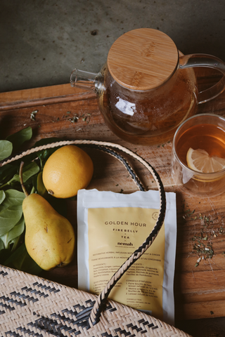 Glass teapot with wooden lid, cup of tea with lemon slice, pear, lemon, woven basket, and a package labeled 'GOLDEN HOUR FIRE BELLY TEA nemah' on a wooden surface.