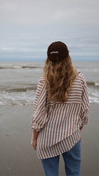 Person wearing a brown hat with the text 'némah' on the back, a beige and white striped shirt, and blue jeans, standing on a beach facing the ocean.