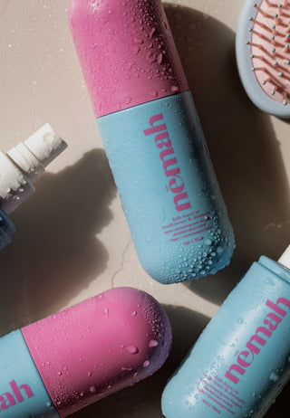Blue and pink nemah shampoo and conditioner bottles wet with water droplets, lying on a beige surface