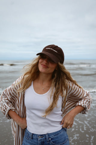 Woman standing on a beach wearing a dark hat with white text, a striped shirt, and a white tank top