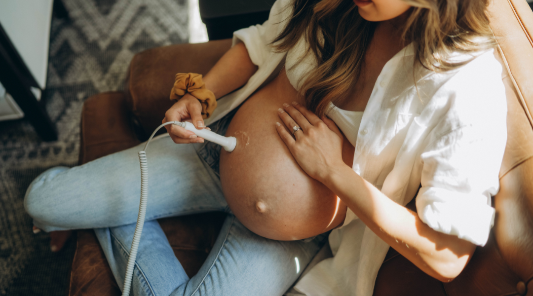 A pregnant woman sitting on a leather couch, using a handheld device to monitor her baby. She is dressed casually in a white shirt and jeans, with natural light illuminating the scene.