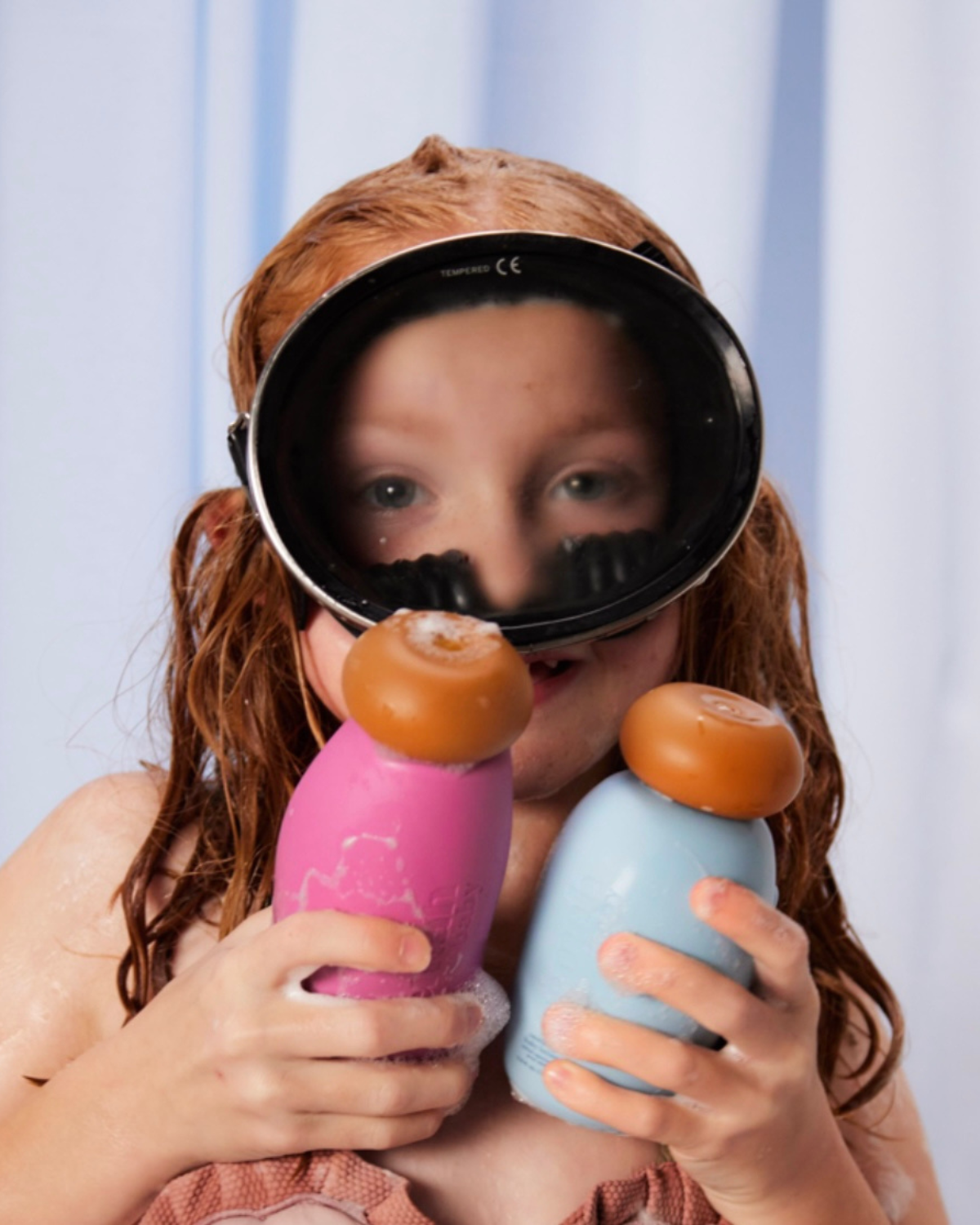 A child in scuba mask is holding in her hands two bottles of ultra gentle baby shampoo and nutrient-rich baby lotion