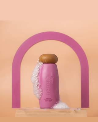 Pink bottle of némah baby nourishing baby wash & shampoo with foam on the side and top, set against a peach background with a purple arch shape behind it.