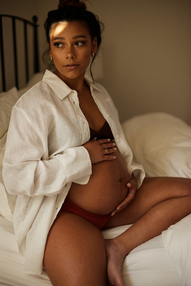 A pregnant woman sitting on a bed, looking thoughtfully to the side. She is wearing a white button-up shirt over red underwear, with sunlight softly lighting the scene through a window.