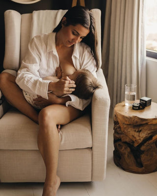 Woman breastfeeding baby while sitting in a beige armchair next to a wooden side table with a glass of water.